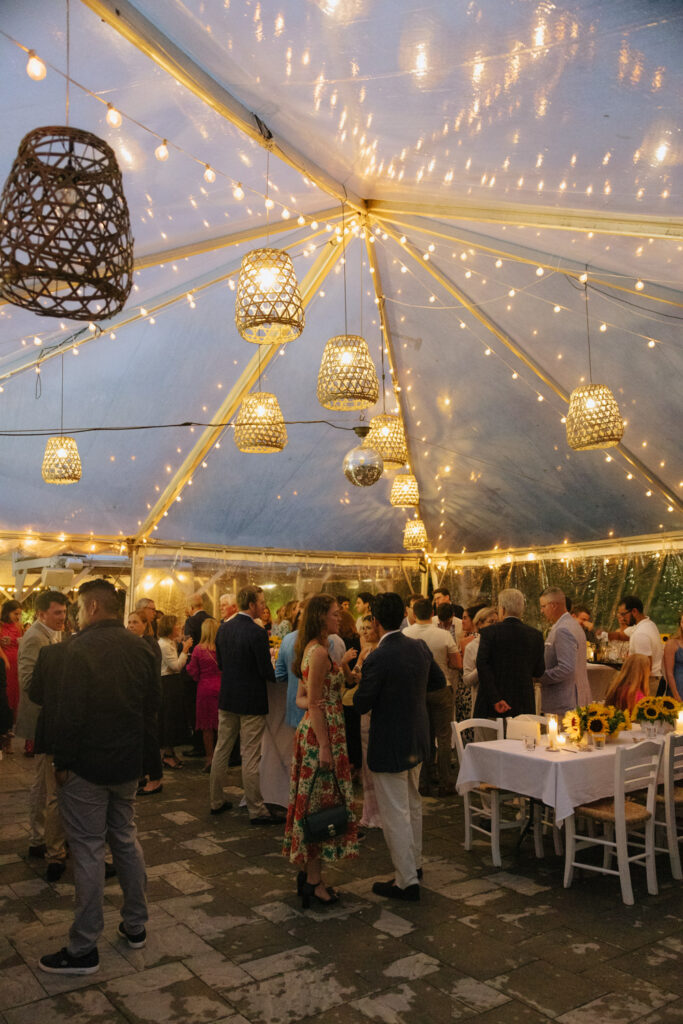 Wedding guests mingle in a clear tent with decorative lighting at Bridgehampton Tennis & Surf Club.