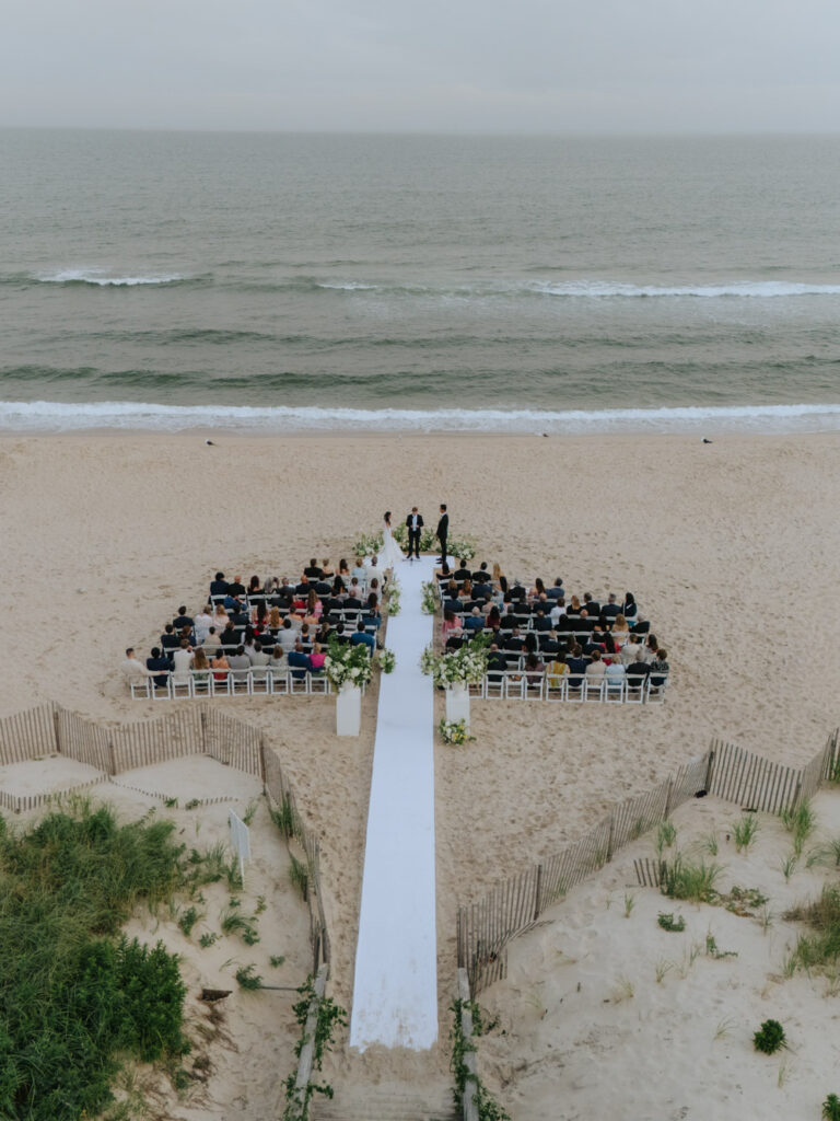 An aerial view of a beachfront wedding ceremony with guests seated and the ocean in the background.