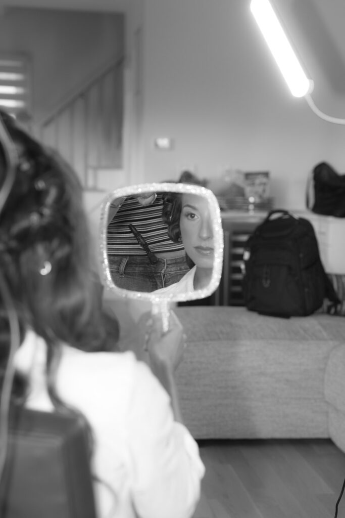 A woman looks into a handheld mirror while getting ready for a Bridgehampton Tennis & Surf Club wedding.