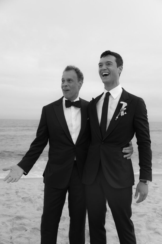 Two men in tuxedos laugh on the beach during a Bridgehampton Tennis & Surf Club wedding.