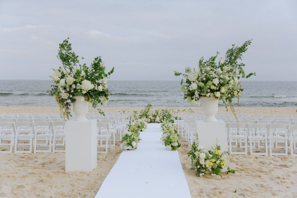Beach wedding ceremony with white chairs, aisle runner, floral arrangements, and the ocean view.