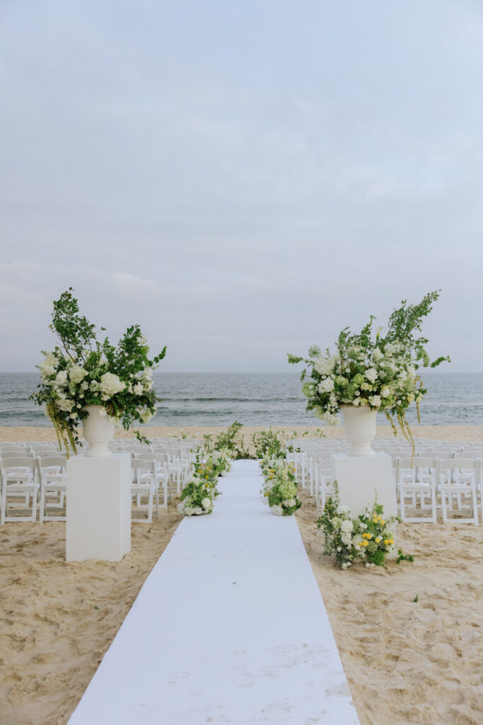An outdoor beach wedding ceremony with white chairs, a white aisle runner, and floral arrangements.