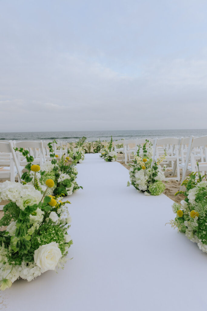 An outdoor beach wedding ceremony setup with white chairs, aisle runner, and floral arrangements.