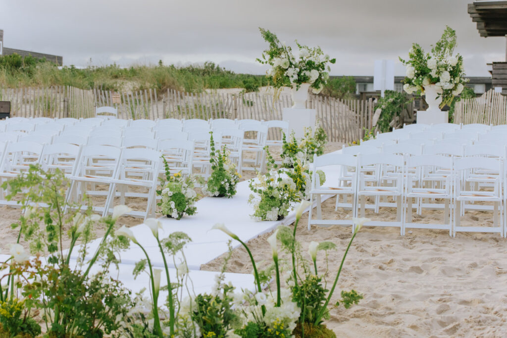 White chairs line an aisle of white carpet and green florals on the sand for a beach wedding at Bridgehampton Tennis & Surf Club.
