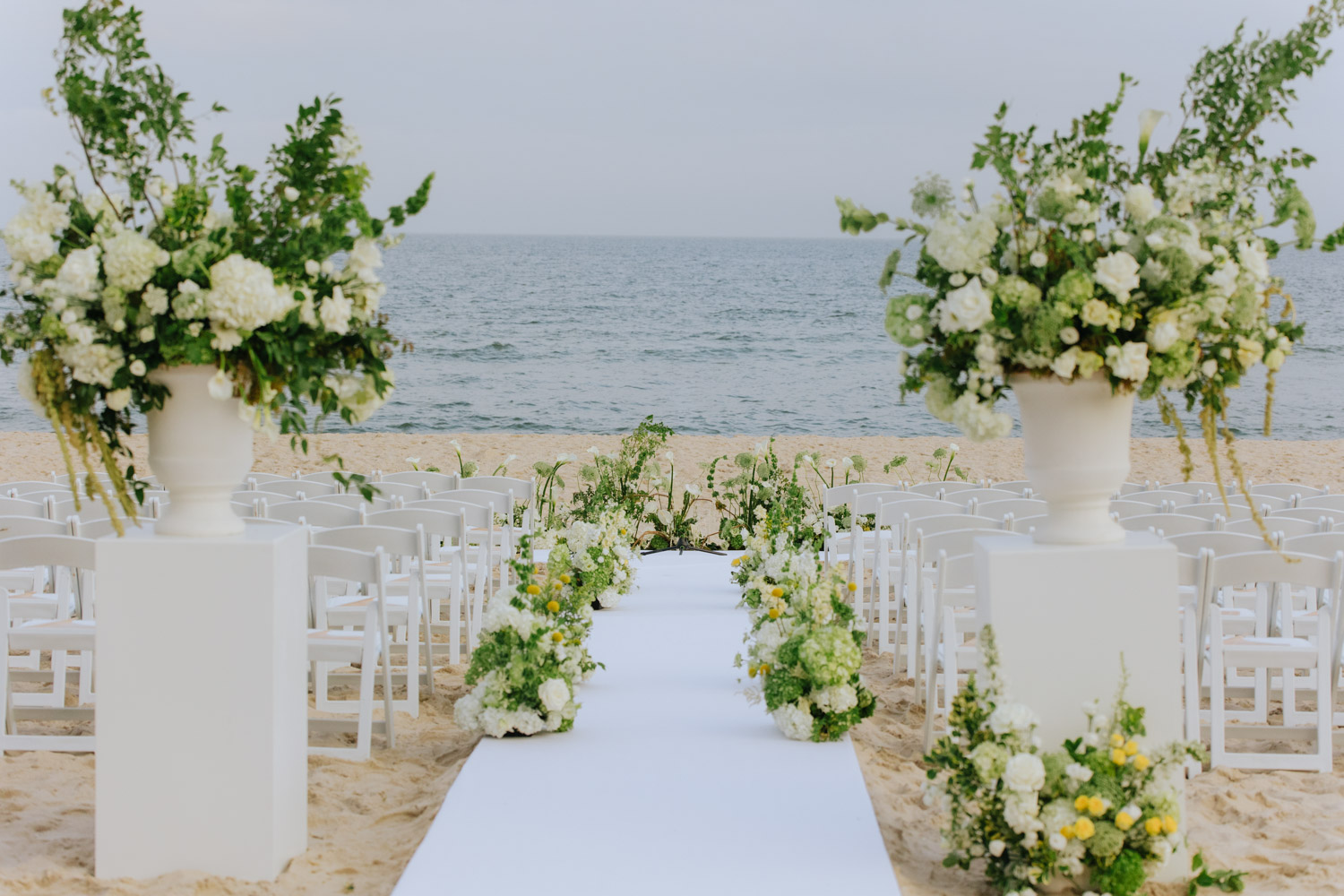 Beach wedding ceremony with white chairs, a white aisle runner, and floral arrangements overlooking the ocean at Bridgehampton Tennis & Surf Club.