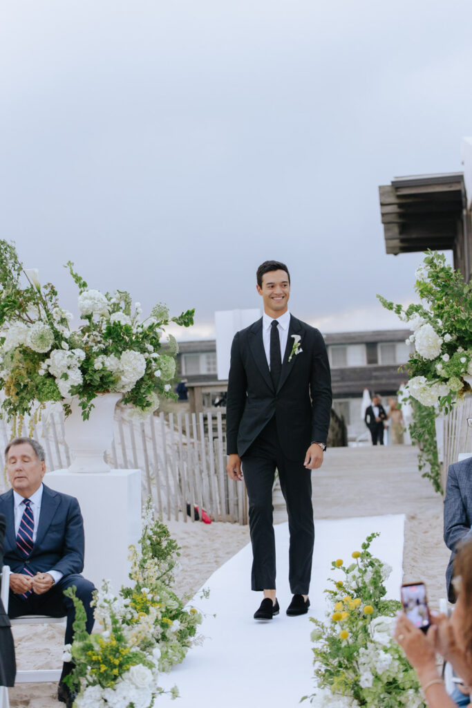 Groom walks down a white aisle on the beach during a Bridgehampton Tennis & Surf Club wedding.