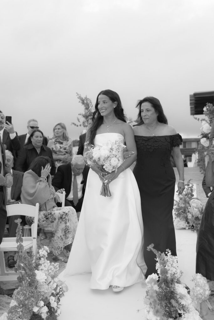 A bride walks down the aisle, holding a bouquet, during a Bridgehampton Tennis & Surf Club wedding.