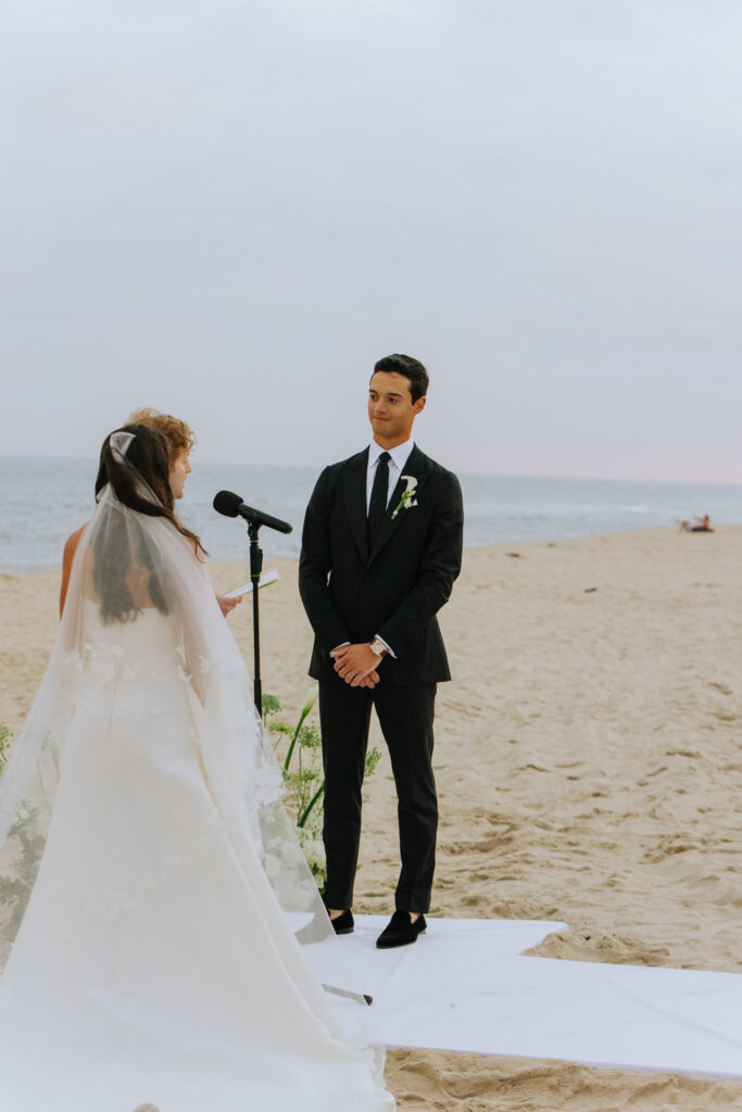 A groom in a black suit looks at his bride during a beach wedding at Bridgehampton Tennis & Surf Club.