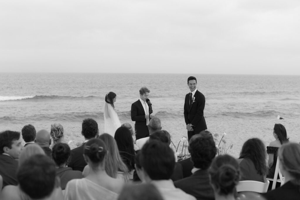A groom smiles as a officiant speaks during a Hamptons beach wedding at Bridgehampton Tennis & Surf Club.