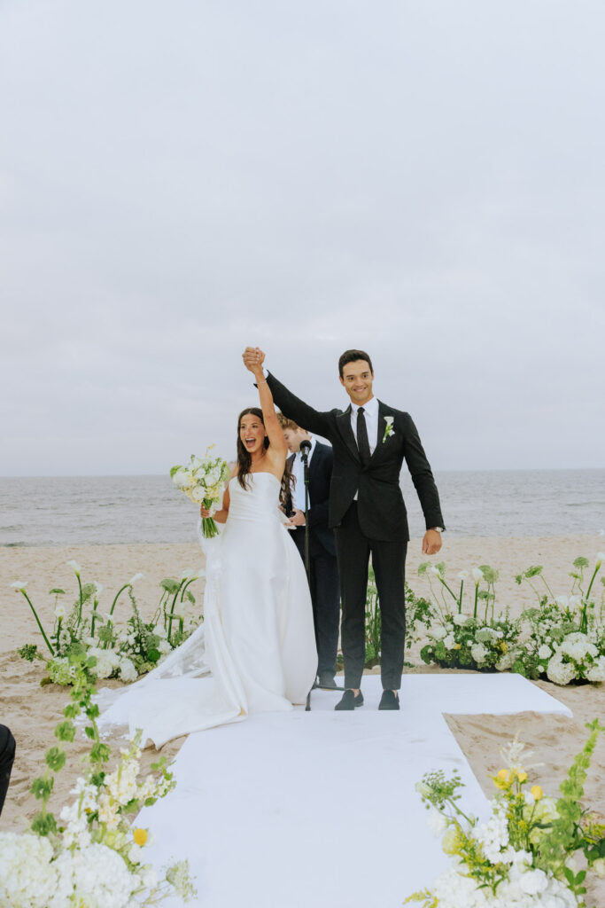 Newlyweds raise their arms in celebration after their beach wedding at Bridgehampton Tennis & Surf Club.