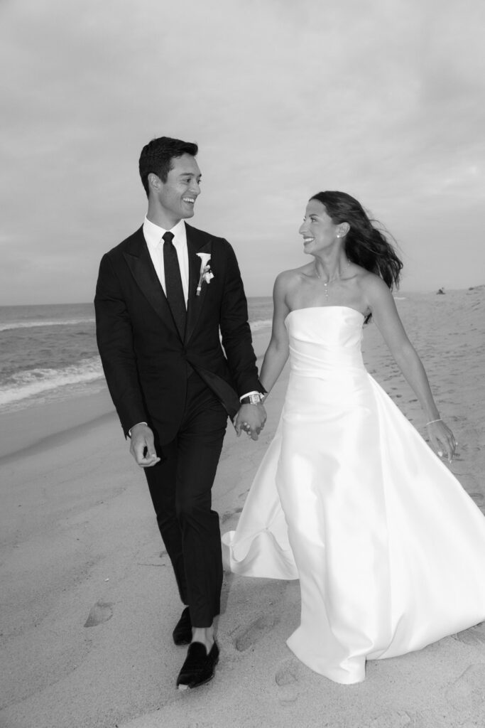 A groom and bride hold hands while walking on the beach at Bridgehampton Tennis & Surf Club.