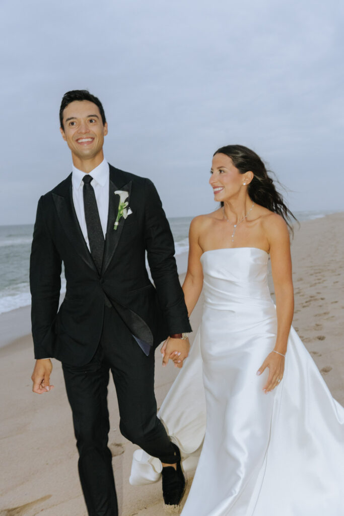 A newlywed couple walks hand in hand on a sandy beach during their Bridgehampton wedding.