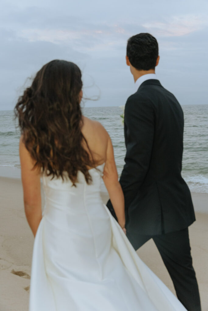 A bride and groom walk hand-in-hand along the Atlantic Ocean beach at their Bridgehampton Tennis & Surf Club wedding.