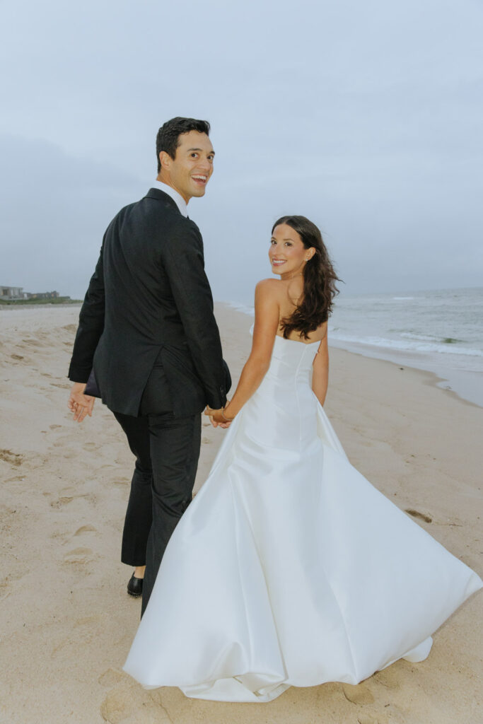 Newlyweds hold hands walking on the beach at their Bridgehampton Tennis & Surf Club wedding.