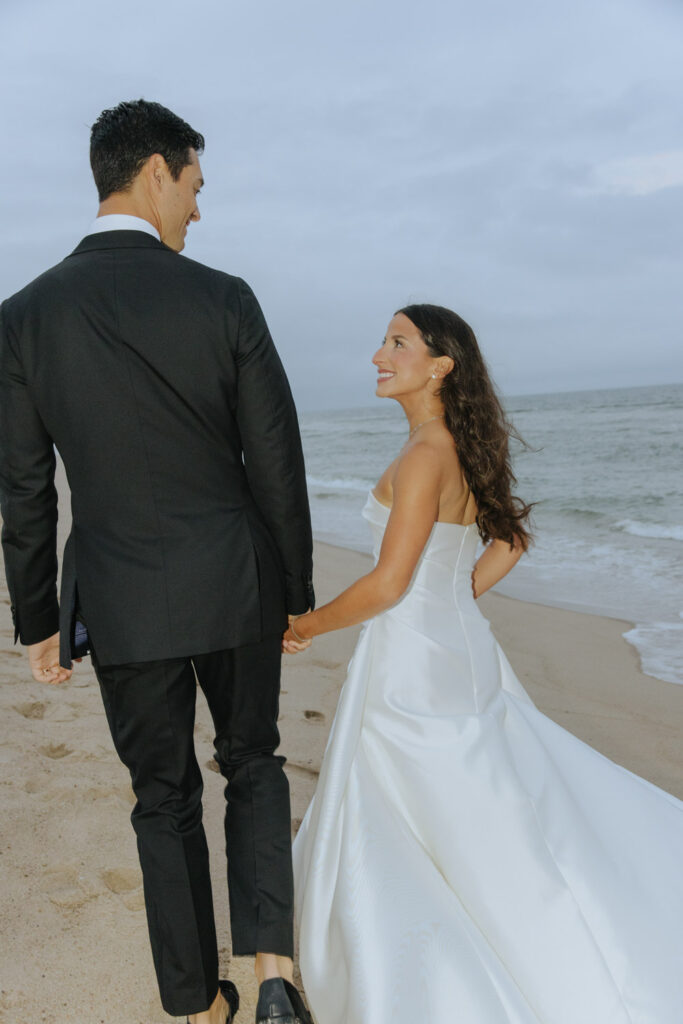 Newlyweds hold hands walking on the sandy beach of their Bridgehampton Tennis & Surf Club wedding.