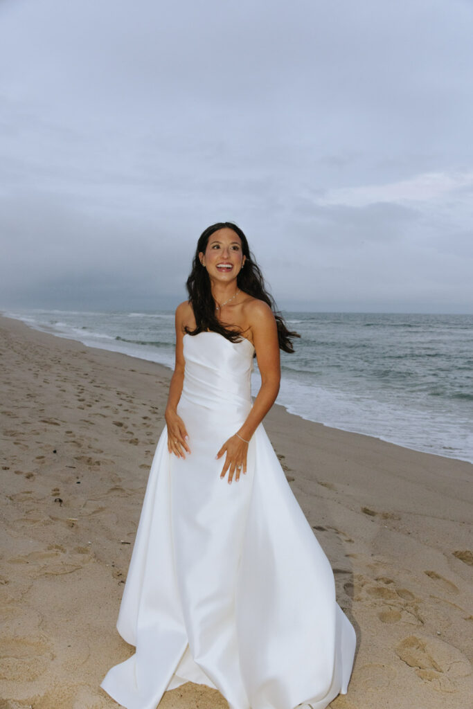 A bride in a white strapless gown smiles on the sand at her Bridgehampton Tennis & Surf Club wedding.