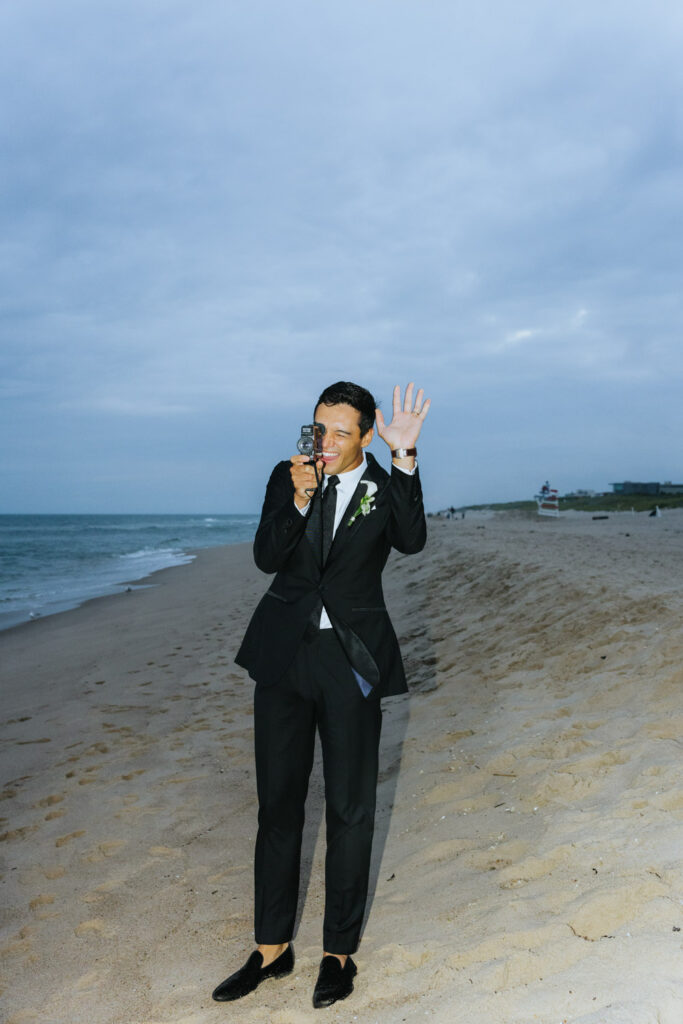 Man waves and holds a camera on the beach during a Bridgehampton Tennis & Surf Club wedding.