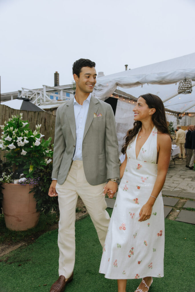 A couple holds hands and smiles, walking past a white tent at a Bridgehampton Tennis & Surf Club wedding.