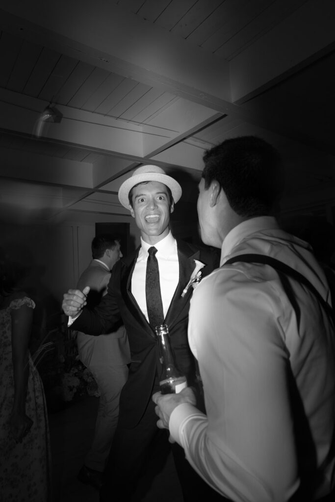 A man in a hat and tuxedo laughs with another man at a Bridgehampton Tennis & Surf Club wedding.