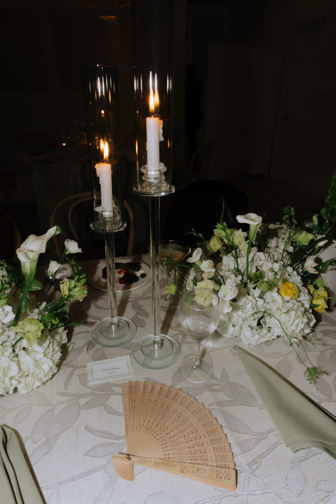 A table setting with lit candles in glass holders, floral centerpieces, and a wooden fan at a Bridgehampton wedding.