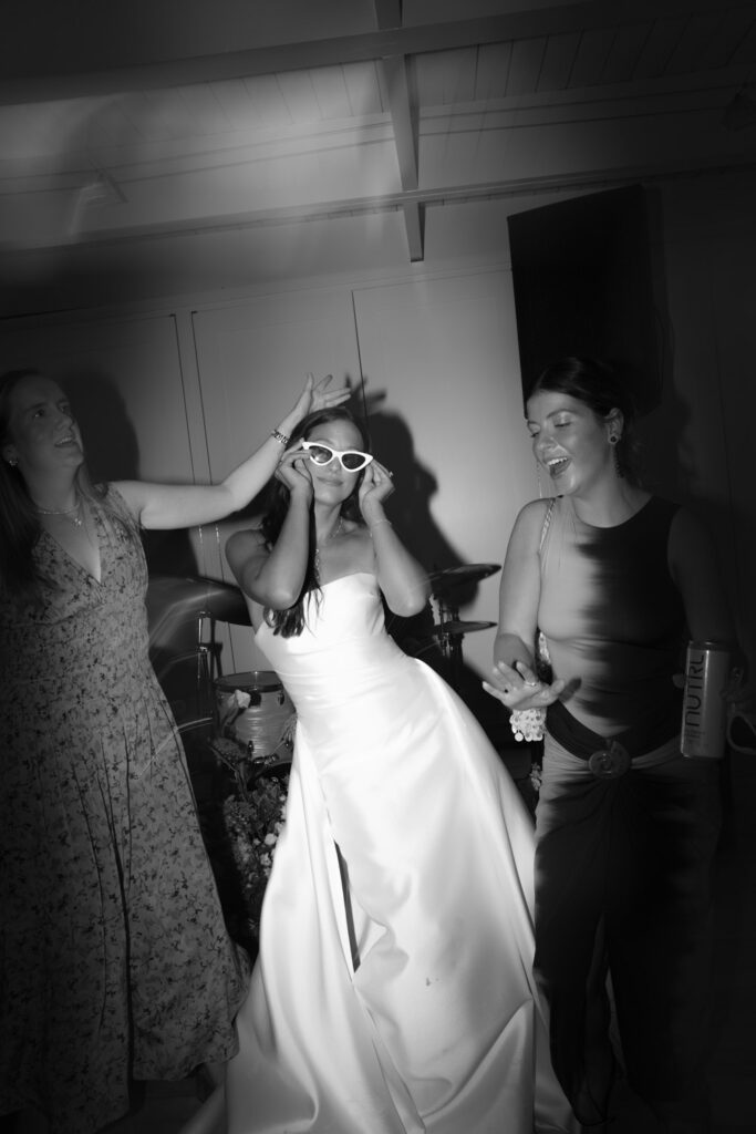 Bride in white gown wearing sunglasses dancing with guests at a Bridgehampton Tennis & Surf Club wedding.