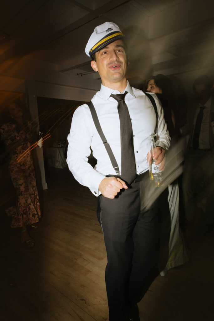Man wearing a captain's hat and suspenders holds a beer during a Bridgehampton Tennis & Surf Club wedding.