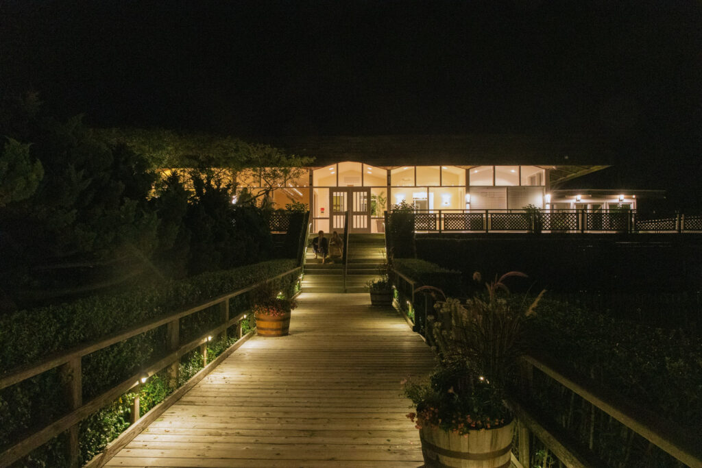 An illuminated wooden walkway leads to the entrance of Bridgehampton Tennis & Surf Club at night.