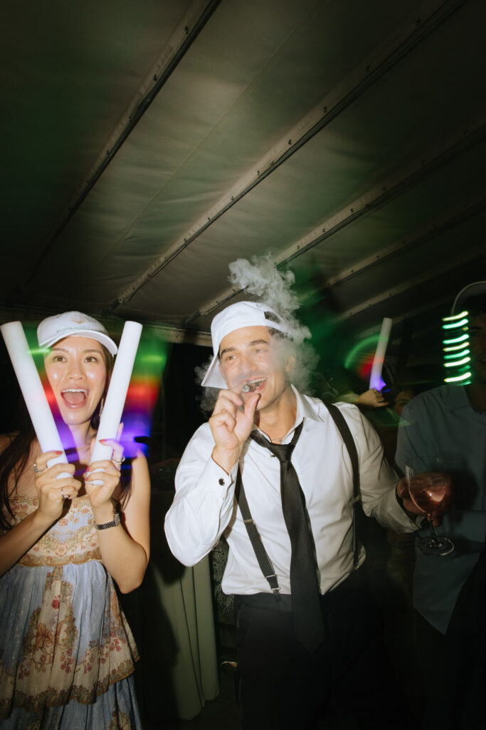 A man smokes a cigar while a woman holds glow sticks at a Bridgehampton Tennis & Surf Club wedding.