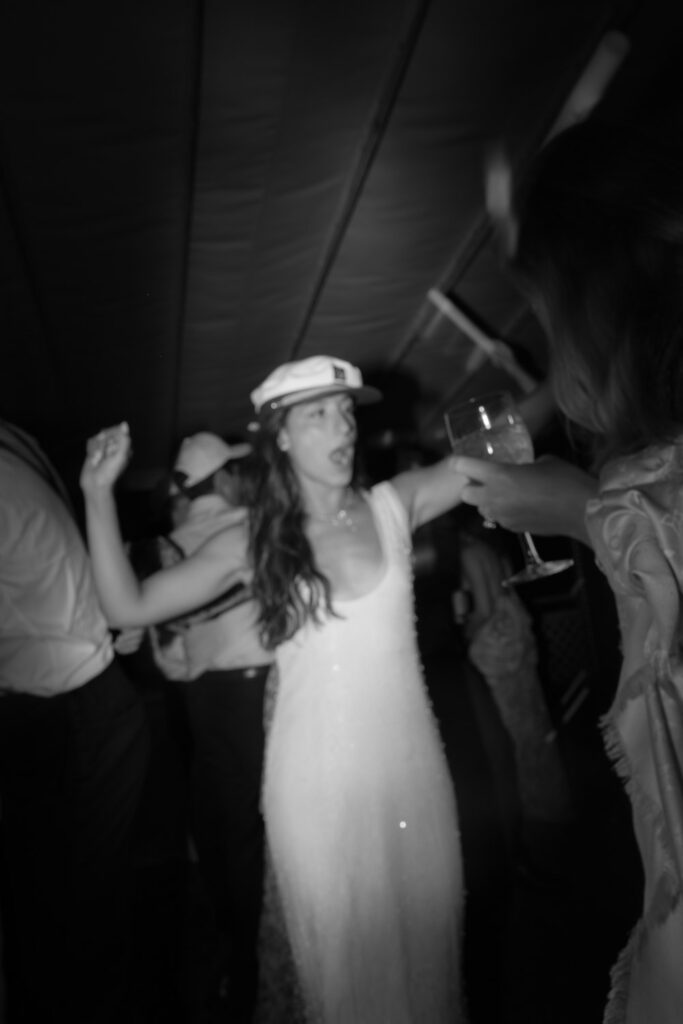 Woman wearing a cap dances with arms raised during a Bridgehampton Tennis & Surf Club wedding.