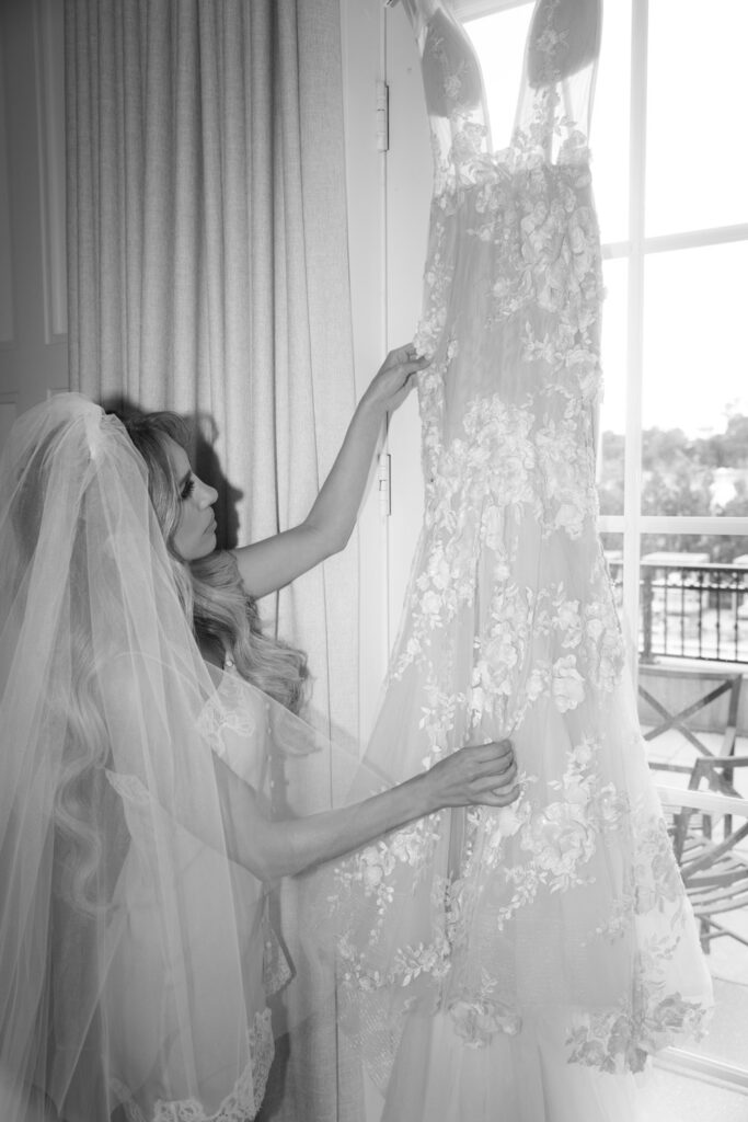 A bride in a veil touches her wedding dress hanging by a window at Russos on the Bay.