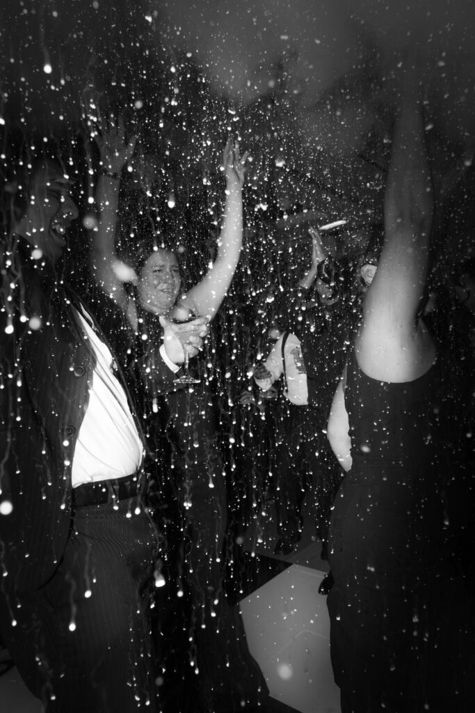 Wedding guests dance on a dark floor with water droplets falling, celebrating a Russos on the Bay wedding.