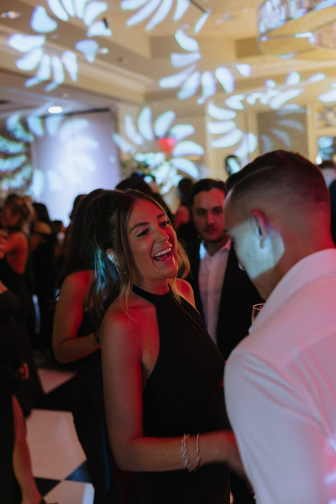 A woman in a black dress laughs while talking to a man at a Russos on the Bay wedding.