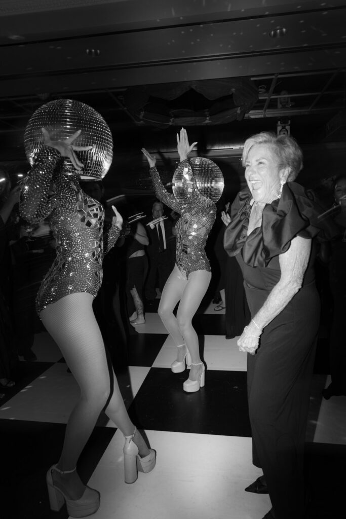 Two performers in disco ball outfits dance with a guest at a Russos on the Bay wedding reception.