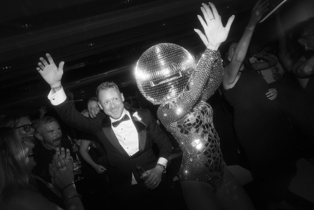 documentary wedding photographer captures A man in a tuxedo smiles while dancing next to a performer in a disco ball costume at a Russos on the Bay wedding.