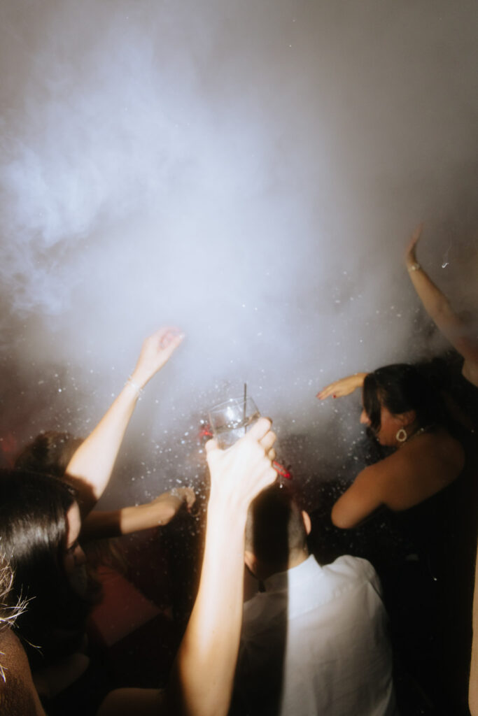 People dancing and raising hands in a smoky, celebratory atmosphere at a Russos on the Bay wedding.