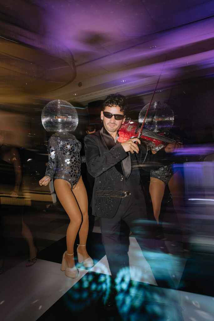A man in sunglasses plays a red violin next to dancers wearing disco ball masks at a Russos on the Bay wedding.