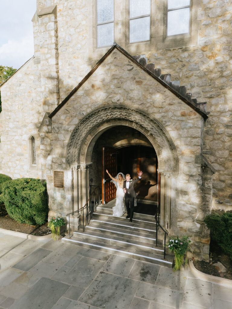The bride and groom wave as they walk down the steps from their Catholic ceremony.