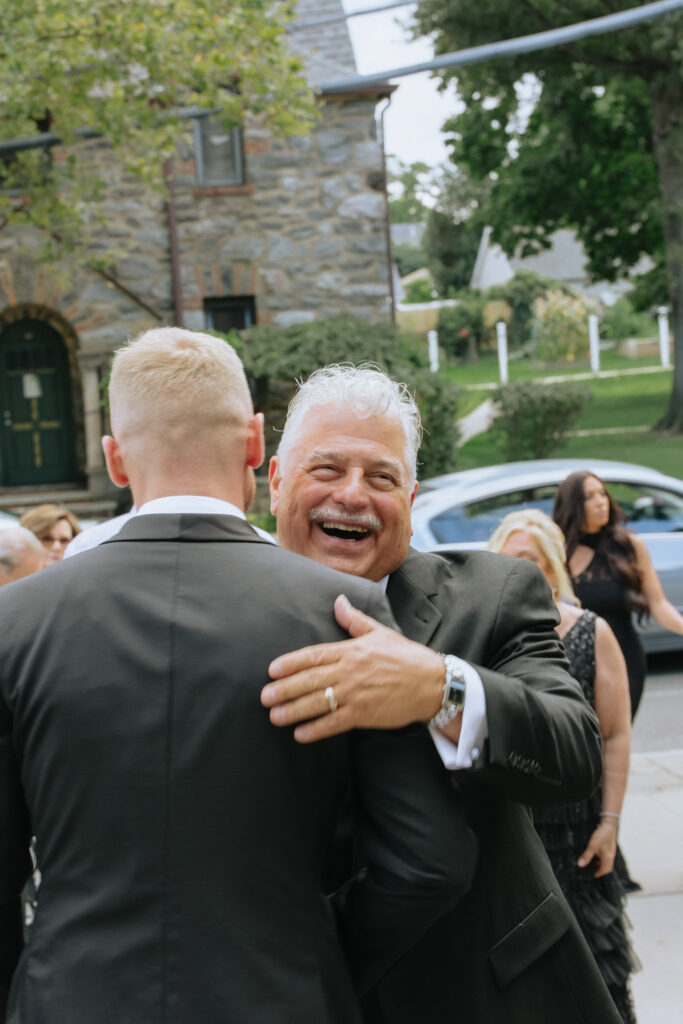 A man in a suit embraces another man, laughing, at a Russos on the Bay wedding reception.