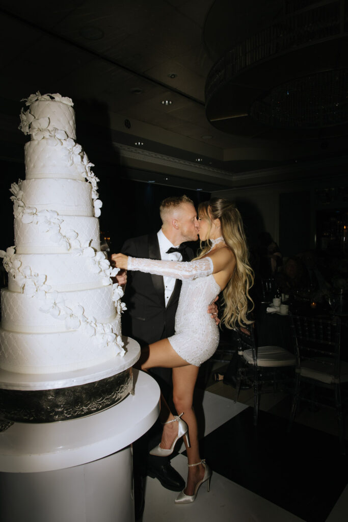 A newlywed couple kisses while cutting their tiered cake at a wedding reception at Russos on the Bay.