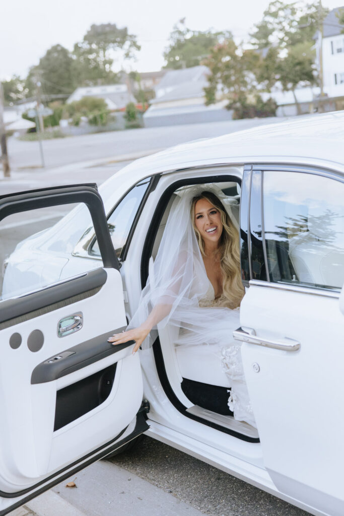 A bride in a white gown and veil smiles as she exits a Rolls-Royce at a Russos on the Bay wedding.
