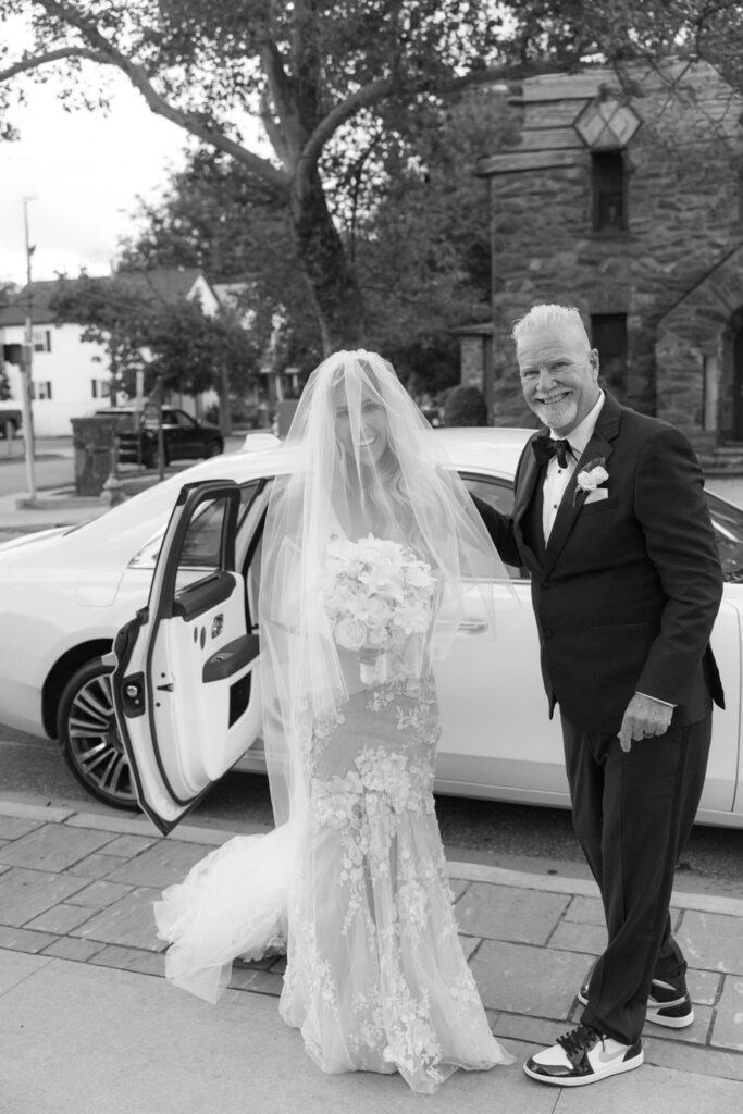 A bride in a white gown and a man in a tuxedo stand next to a Rolls-Royce at a Russos on the Bay wedding.