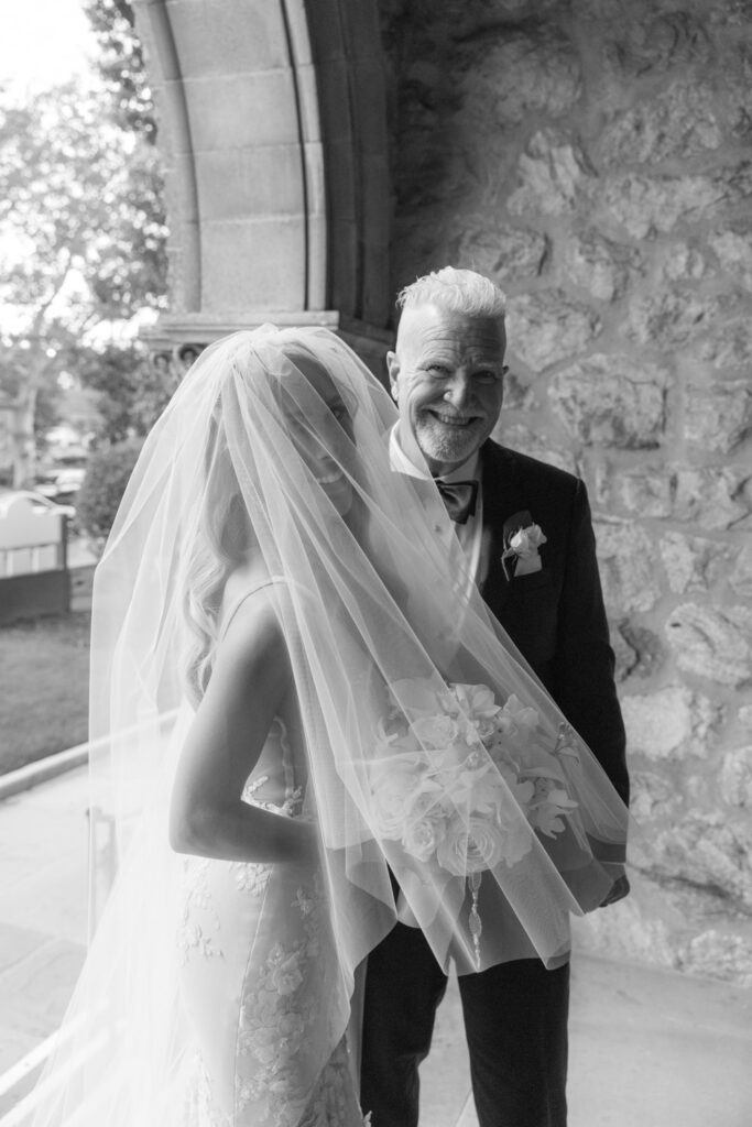 A smiling man standing behind a bride in a white dress and veil at a Russos on the Bay wedding.