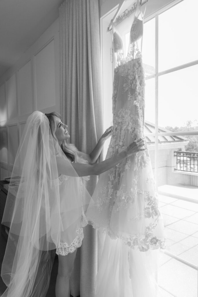 A bride in a veil and lingerie gazes at her wedding dress hanging by a window at Russos on the Bay.
