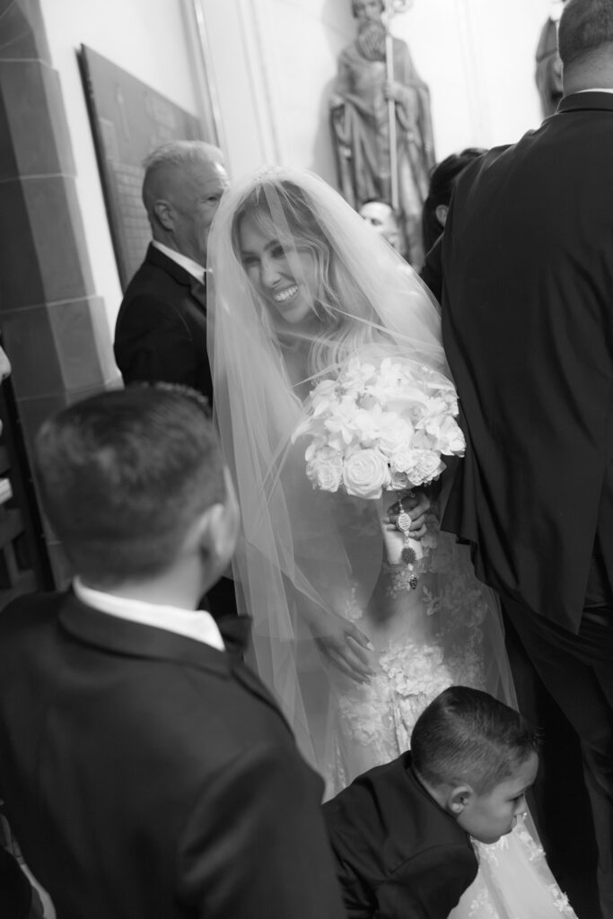 A bride in a white wedding gown, veil, and white bouquet smiles during a Catholic ceremony at Russos on the Bay.