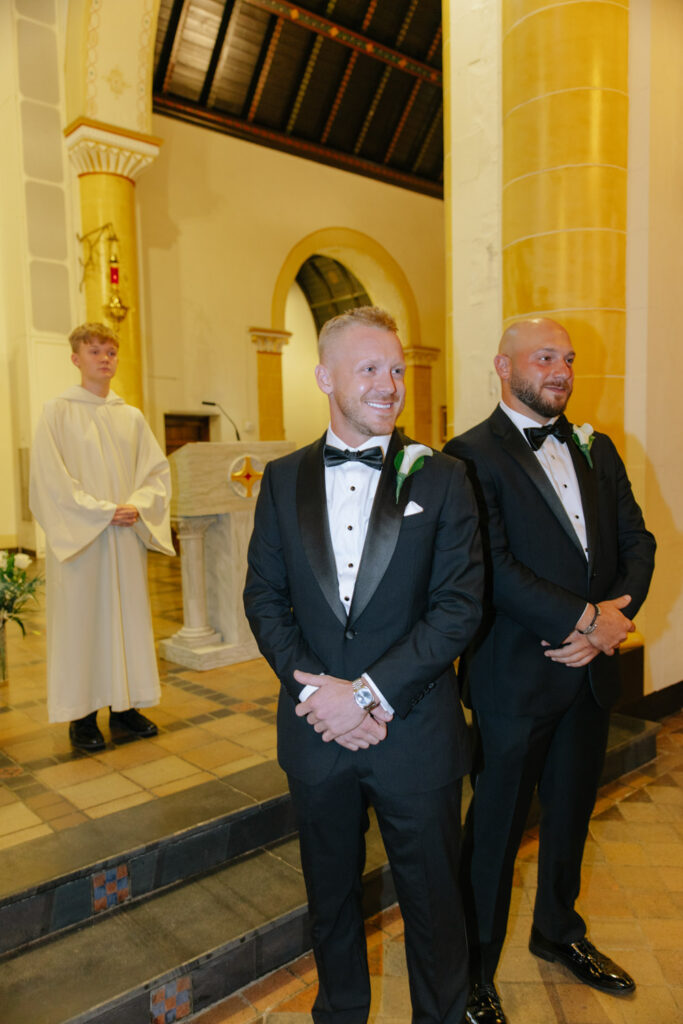 Two men in tuxedos stand at a Catholic wedding ceremony at Russos on the Bay.