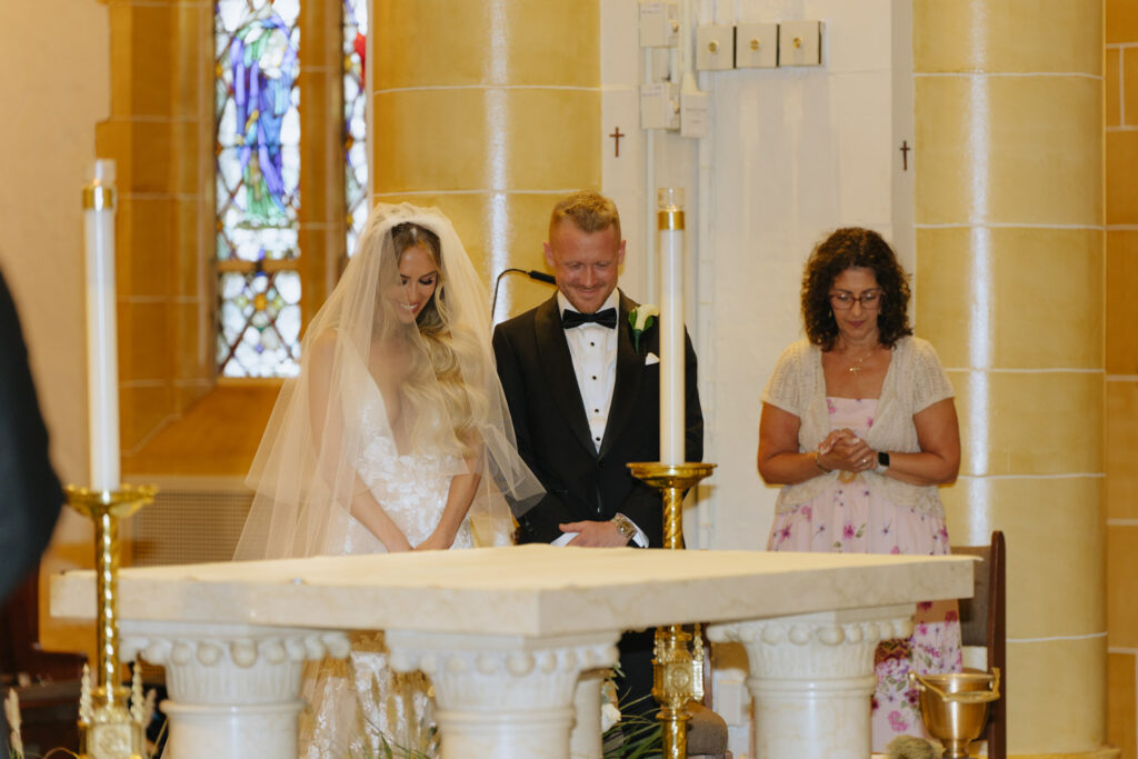 A bride, groom, and guest stand during a Catholic wedding ceremony at Russos on the Bay.