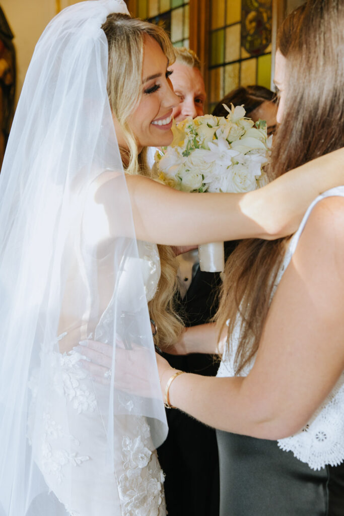 A blonde bride in a white veil smiles, holding a white bouquet, as two women embrace her at a wedding.
