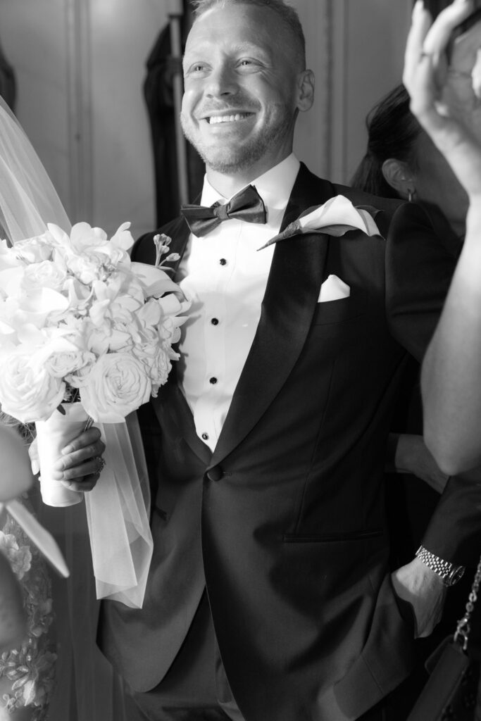 A groom in a tuxedo smiles while holding a wedding bouquet at a Russos on the Bay wedding.