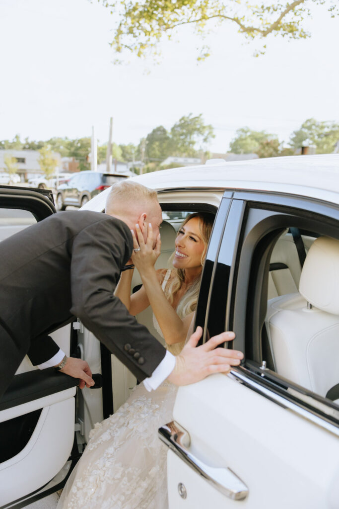 A groom in a suit leans into a white Rolls-Royce, touching the bride's face at Russos on the Bay.