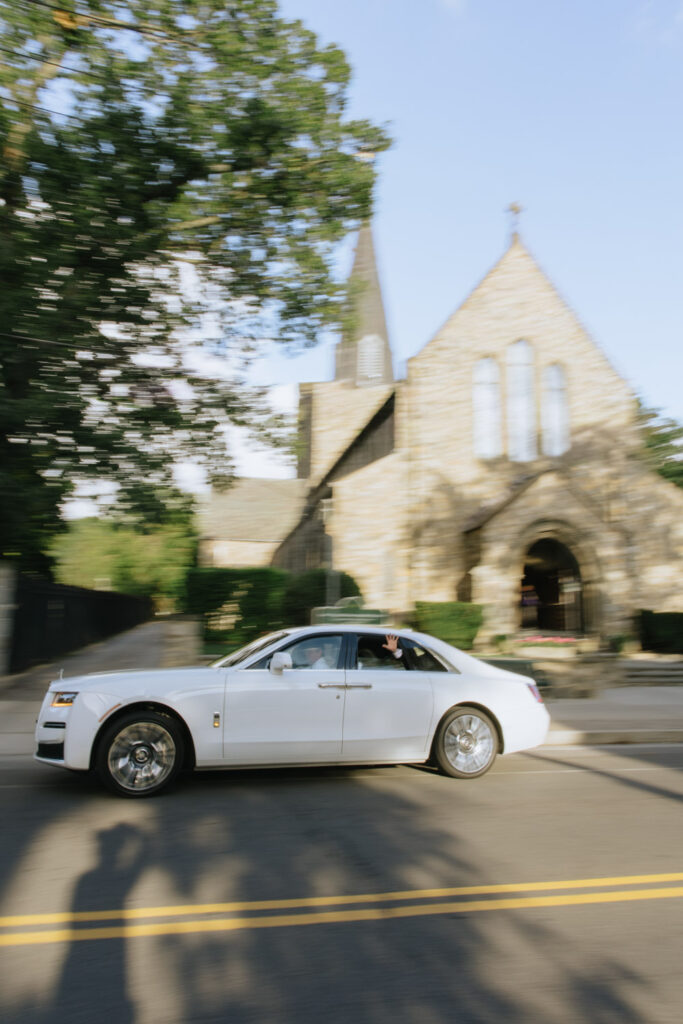 A white Rolls-Royce drives past a church, with a hand waving from the back window, at a Russos on the Bay wedding.